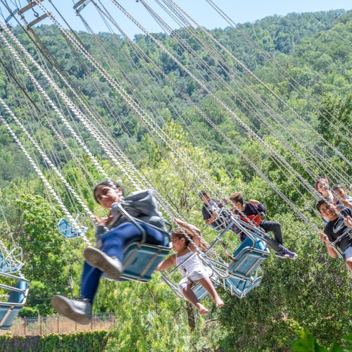 Orang ramai menikmati permainan buaian di taman hiburan dengan latar belakang pokok-pokok hijau yang subur dan bukit-bukau.
