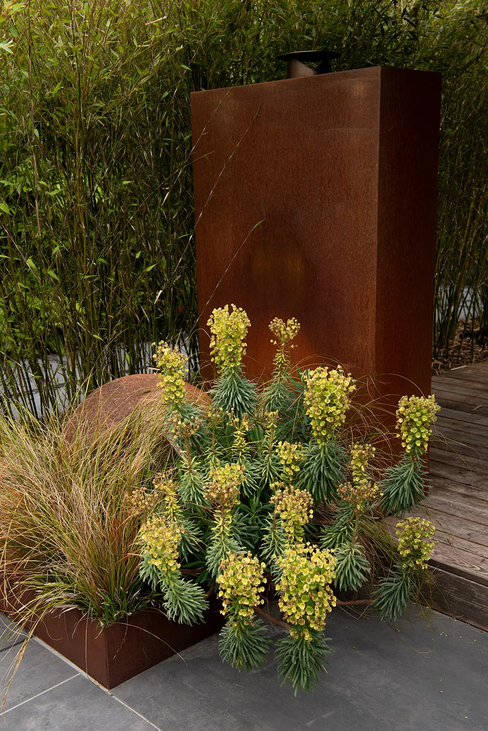 A modern garden scene featuring yellow flowers in front of a rust-colored metal panel and lush greenery.