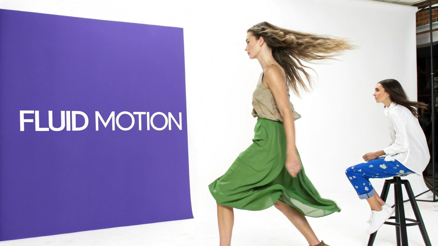 Two fashion models on a studio set, one walking with flowing hair and skirt, the other seated.