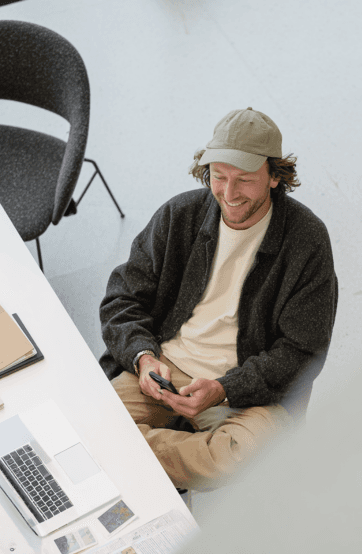 A person in a hat sits at a table, focused on their smartphone, with chairs visible around them.