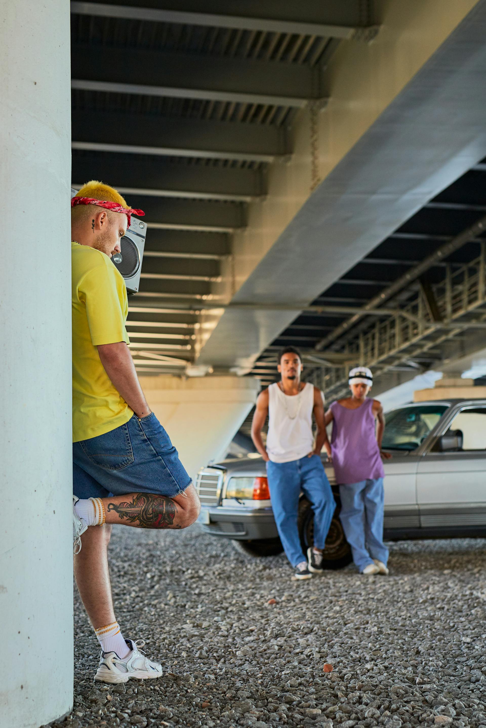 A man with yellow hair and a red bandana leans against a concrete pillar with a boombox on his shoulder, while two other men stand by a car in the background.