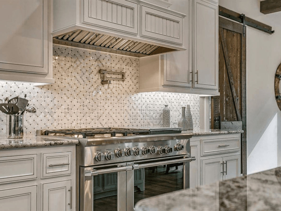 Gas range under a custom vent hood with a tile backsplash and white cabinetry