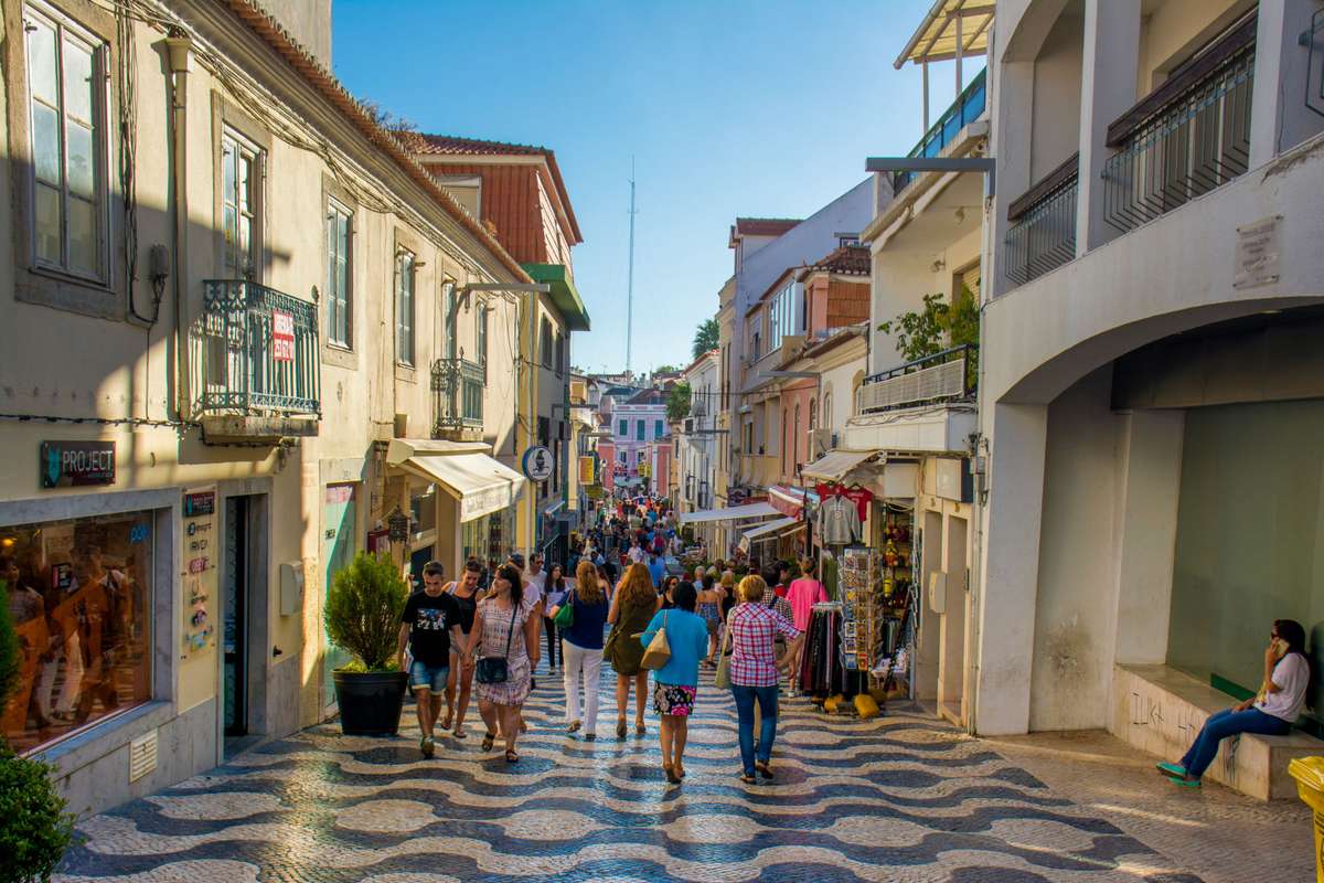 Crowds of people walk along a narrow cobblestone street lined with white buildings and shops in a coastal village.