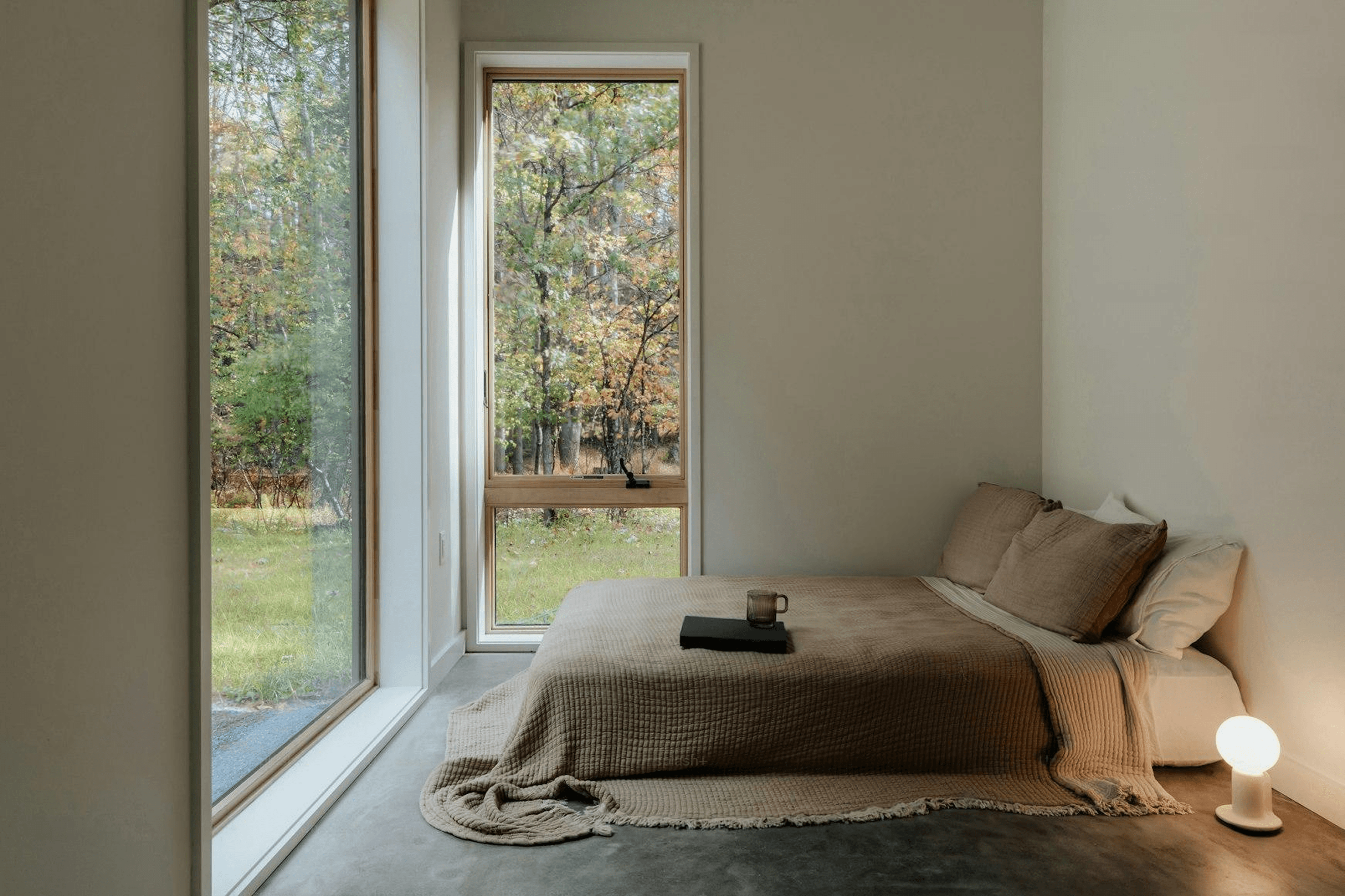 Interior of a Strawtown Stack One modular home bedroom with a tan waffle-weave duvet, concrete floor, and large window overlooking a green forest