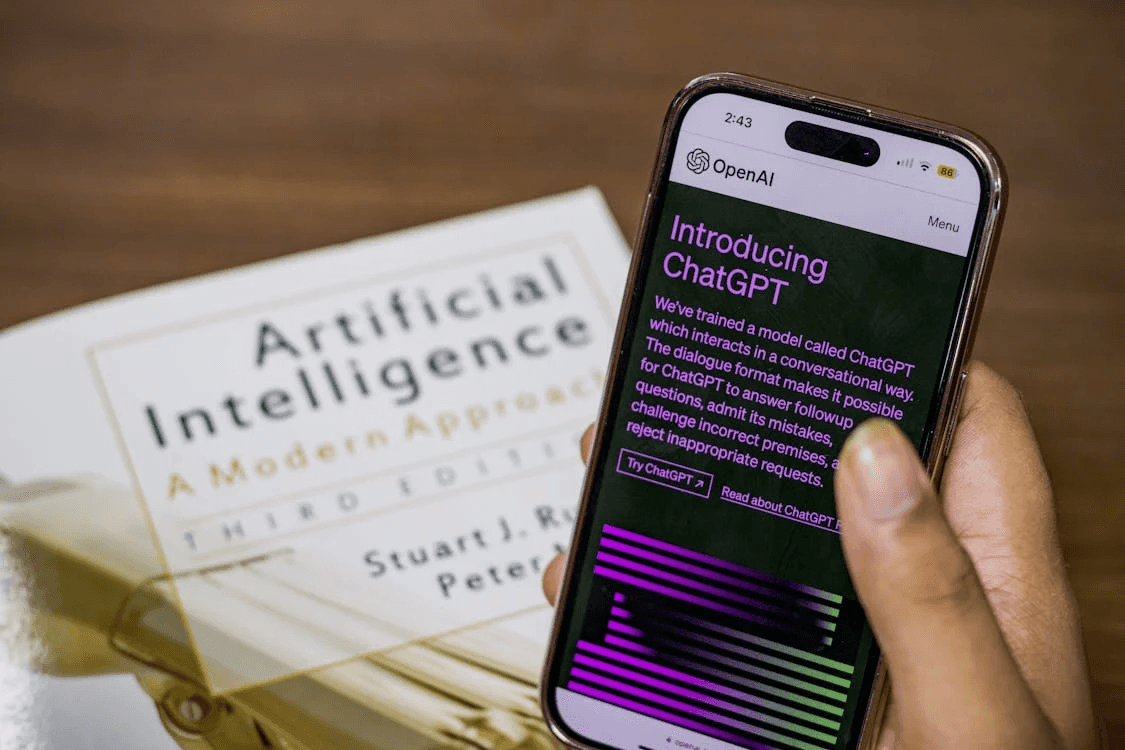 A hand holding a smartphone showing an OpenAI “Introducing ChatGPT” page, resting over the book Artificial Intelligence: A Modern Approach on a desk.