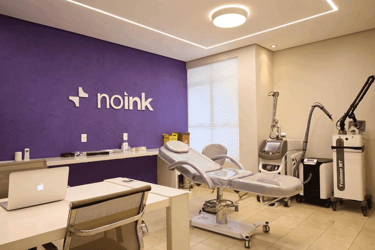 Consultório da clínica NO INK com equipamentos modernos de remoção de tatuagem a laser, maca estética branca e parede de destaque roxa.
