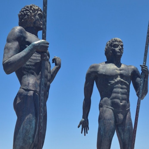 两座青铜雕像，分别为肌肉发达的古代战士形象，手持长矛，背景是一片清澈的蓝天。