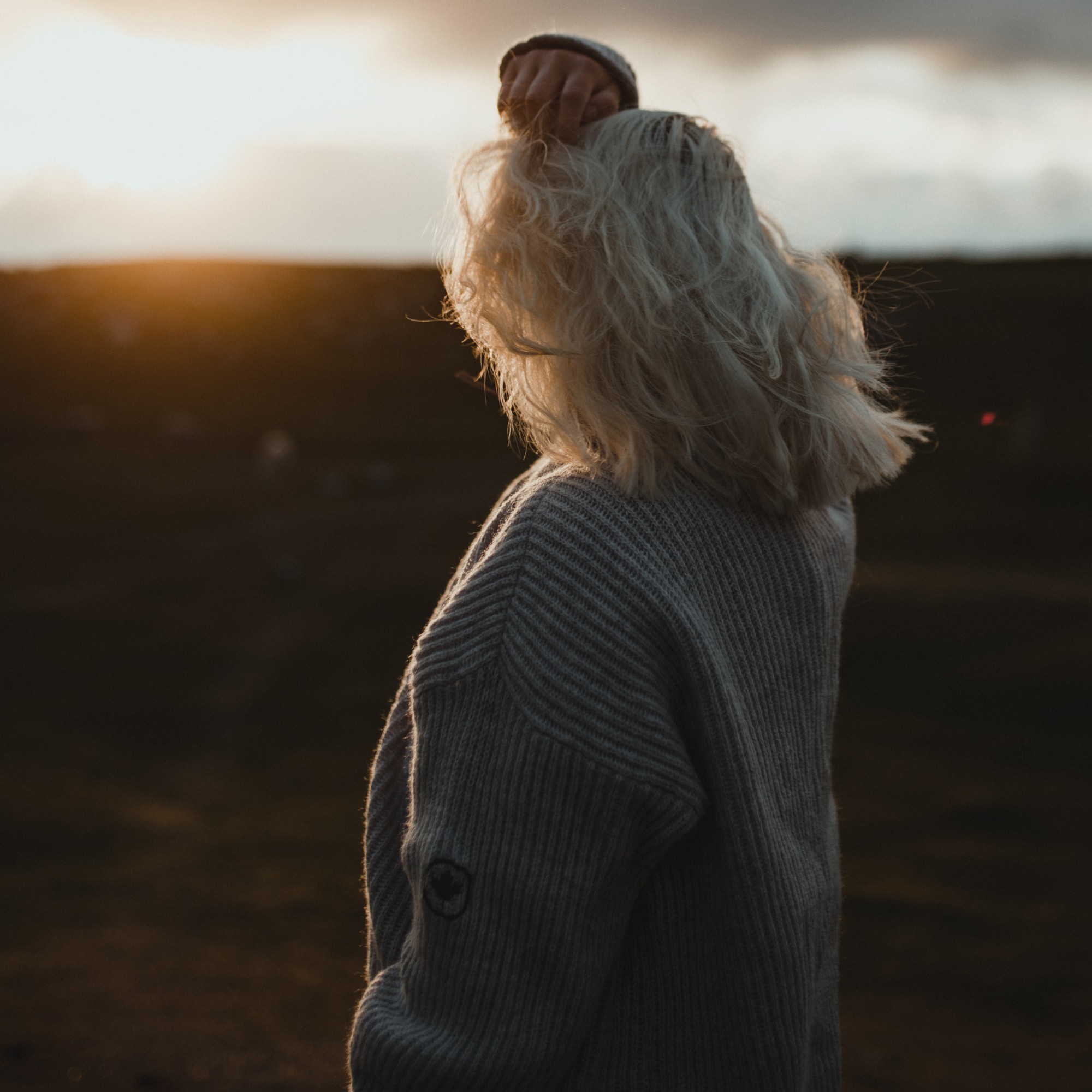 Woman with platinum blonde hair wearing wool sweater, standing in nature at sunset.
