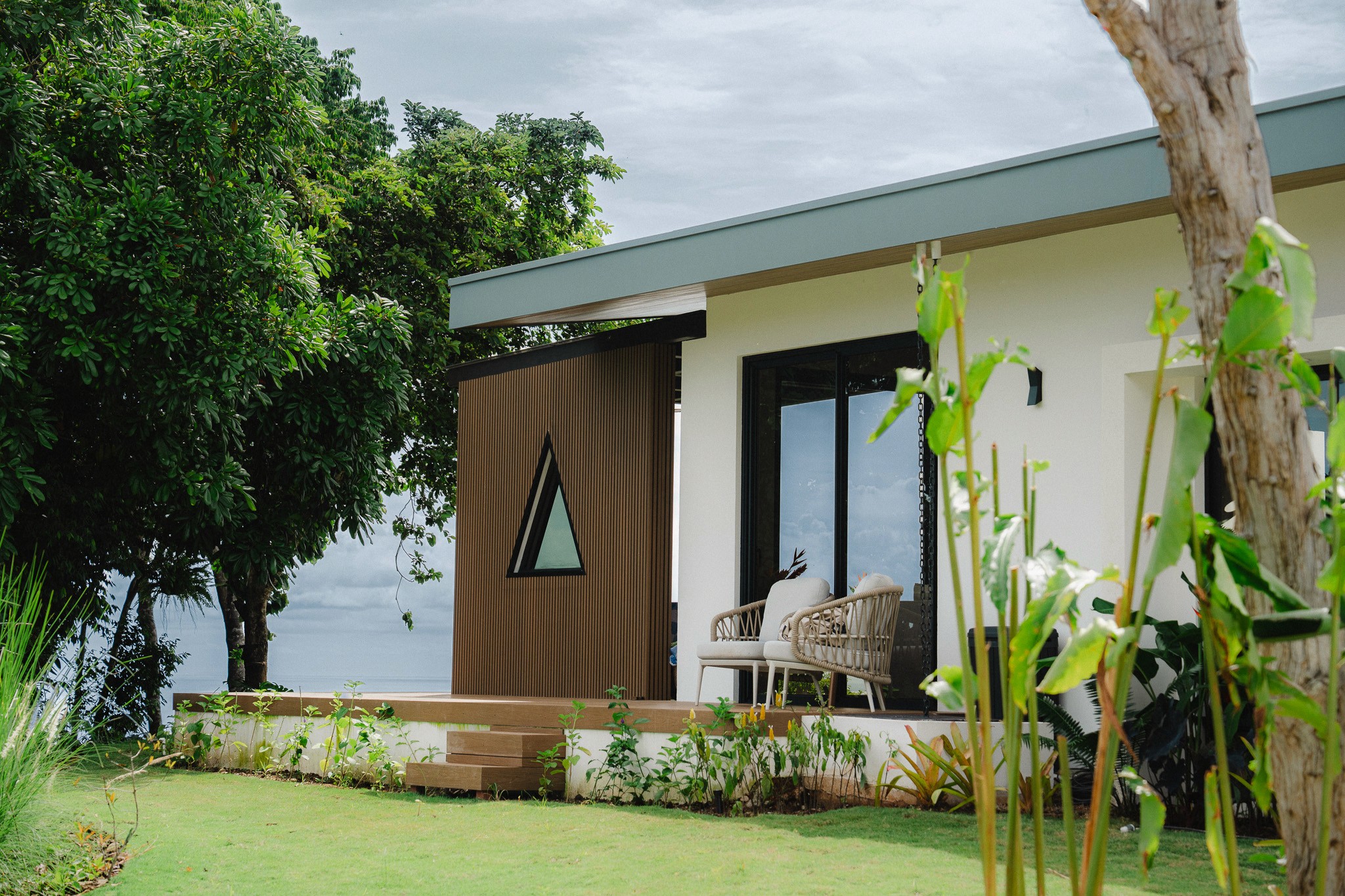 Exterior view of a private deck seating area on a modern Costa Rica villa, featuring unique wood siding and a triangular window nestled among tropical trees.