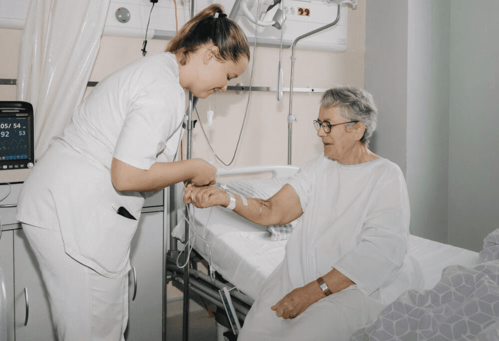 A healthcare provider in white coat examining an elderly patient with glasses in a hospital room, with medical monitoring equipment visible in the background