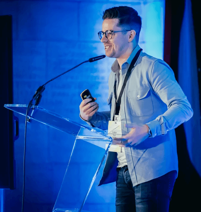 A person stands at a clear lectern on a stage, speaking into a microphone while holding a small handheld device. A large screen to the left displays partially visible presentation text. The setting is lit with blue stage lighting, and the speaker wears a light button-up shirt and a lanyard with a name badge.