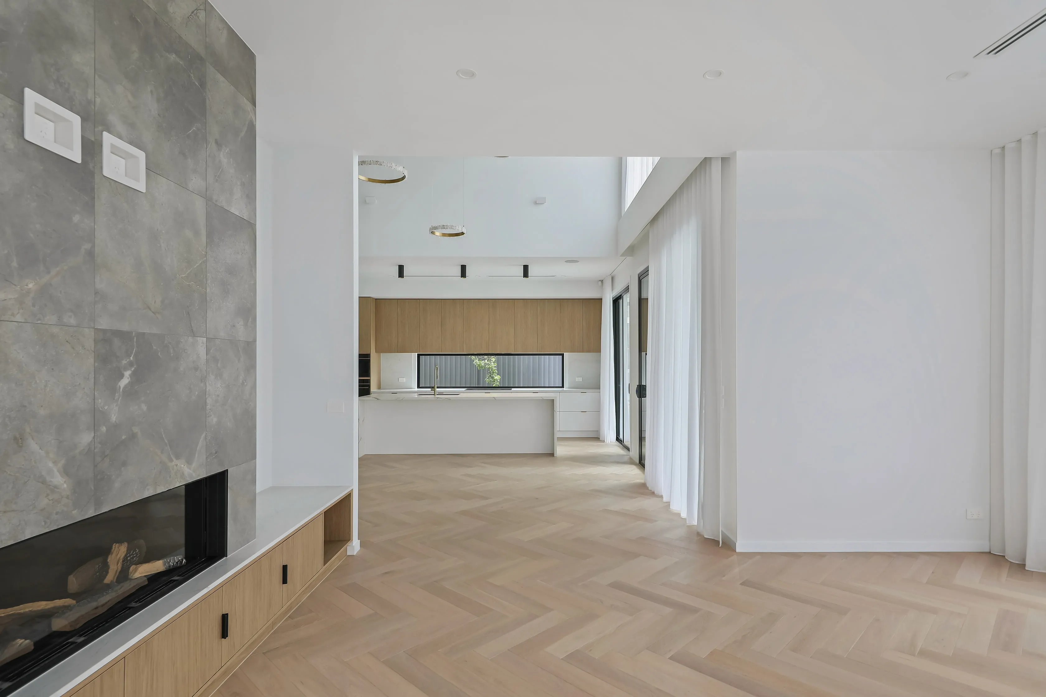 Living and kitchen space at Mount Waverley Residence showing the stone fireplace, double-height void with ring pendant lights and herringbone floors