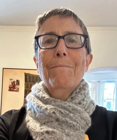 Uma pessoa de cabelo curto, usando óculos e um cachecol de tricô cinza, posa em ambiente interno, com um quadro na parede e a suave luz do dia entrando por uma janela ao fundo.