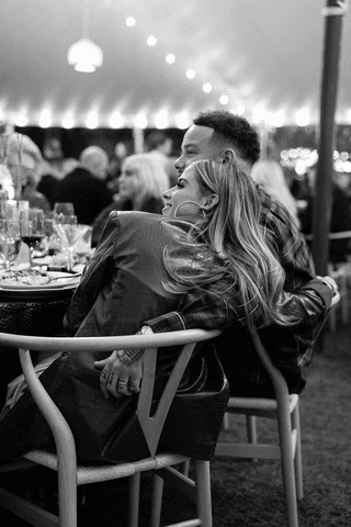 A person with shoulder-length blonde hair wearing a black newsboy cap and a black sweater.