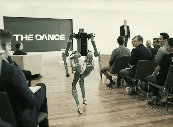 A bipedal robot walks down an aisle toward an audience seated in a modern presentation room with "THE DANCE" displayed on a large screen.
