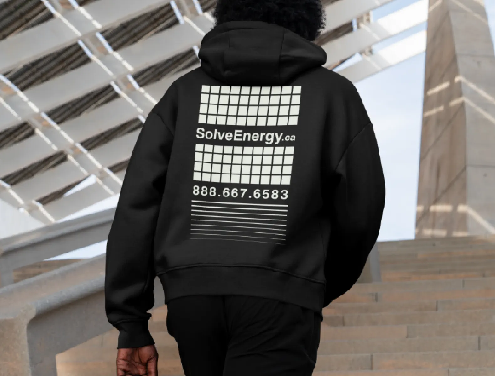 A man walking up stairs wearing a Solve Energy hoodie. The hoodie has solar grids, cascading ines, solveenergy.ca and a phone number printed on it.