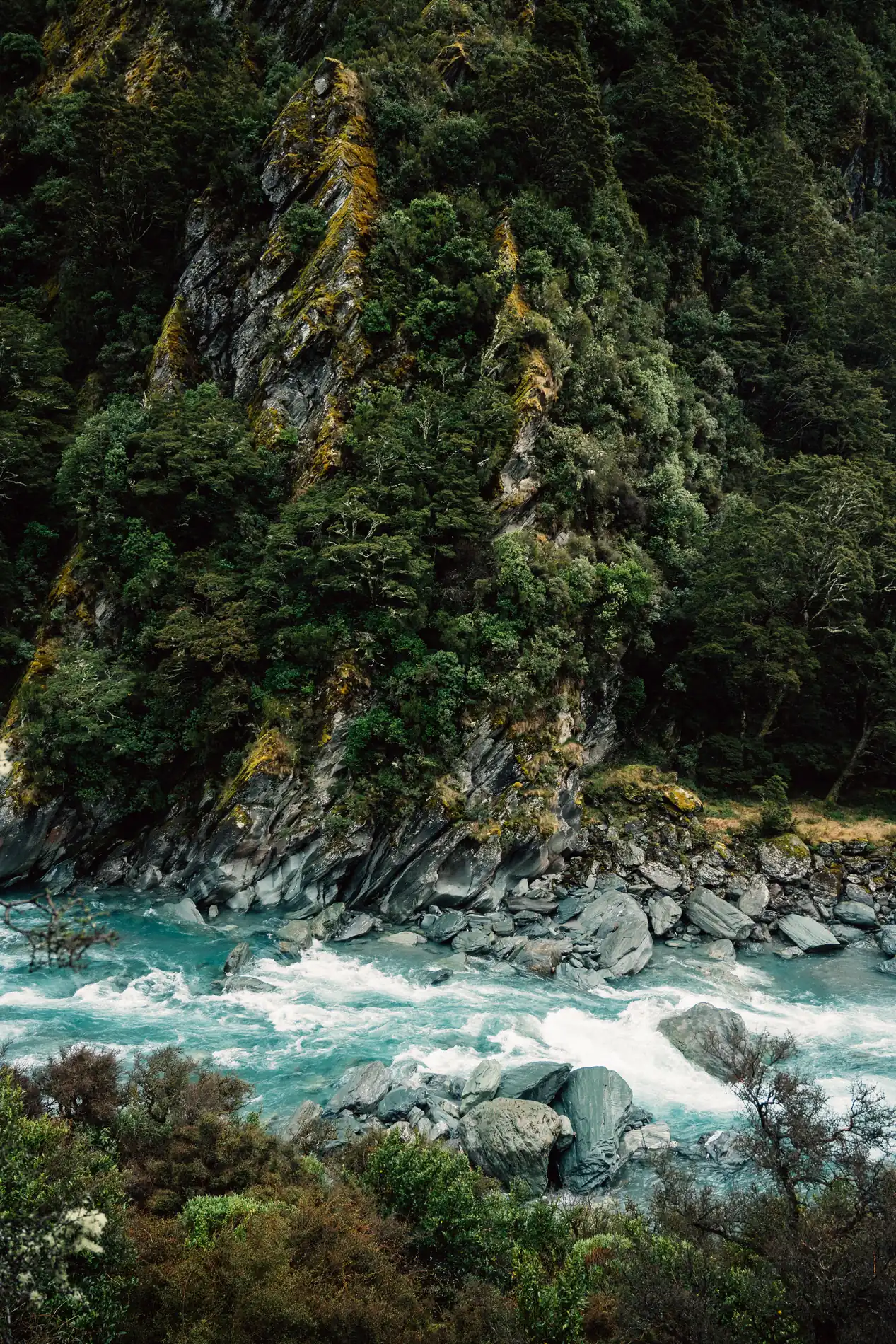 A fast river running over pale boulders below steep, bush-covered rock faces