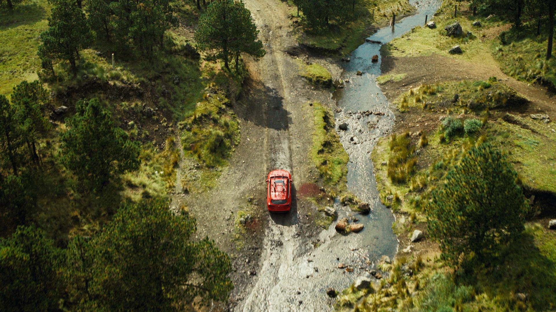 Aerial view of a red SUV driving along a dirt road and crossing a shallow stream through a green, wooded landscape.