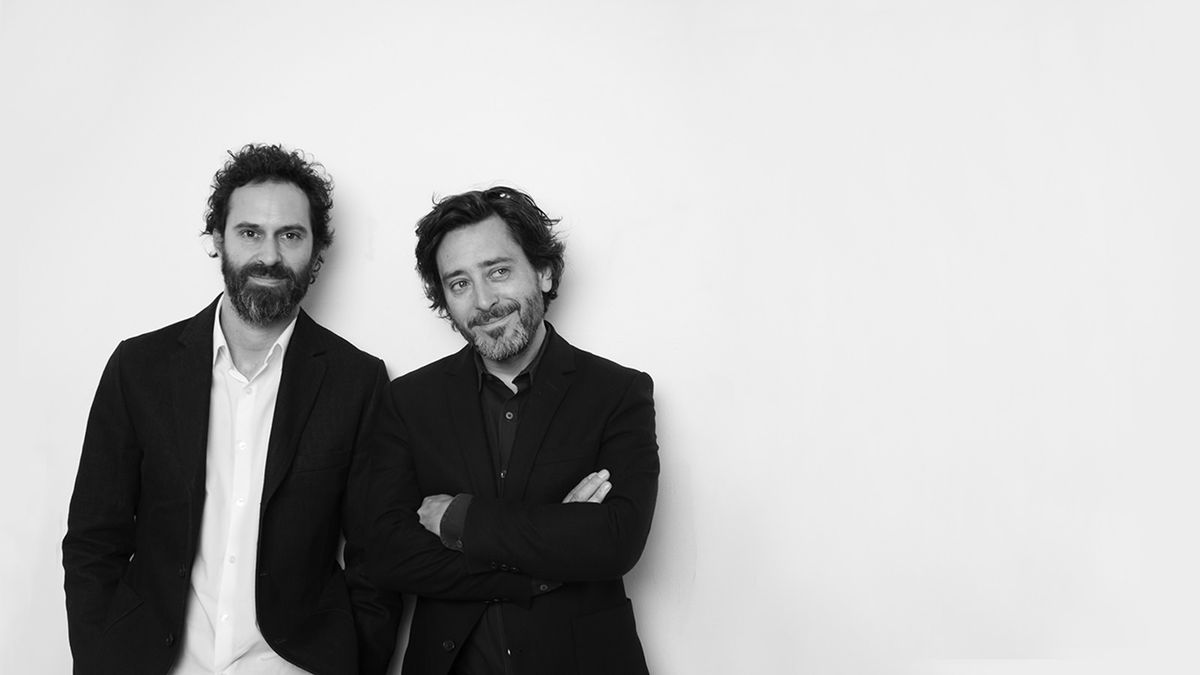 A man in a blue bomber jacket stands against a dark background. The lighting is dramatic, highlighting his focused expression. His pose is confident.