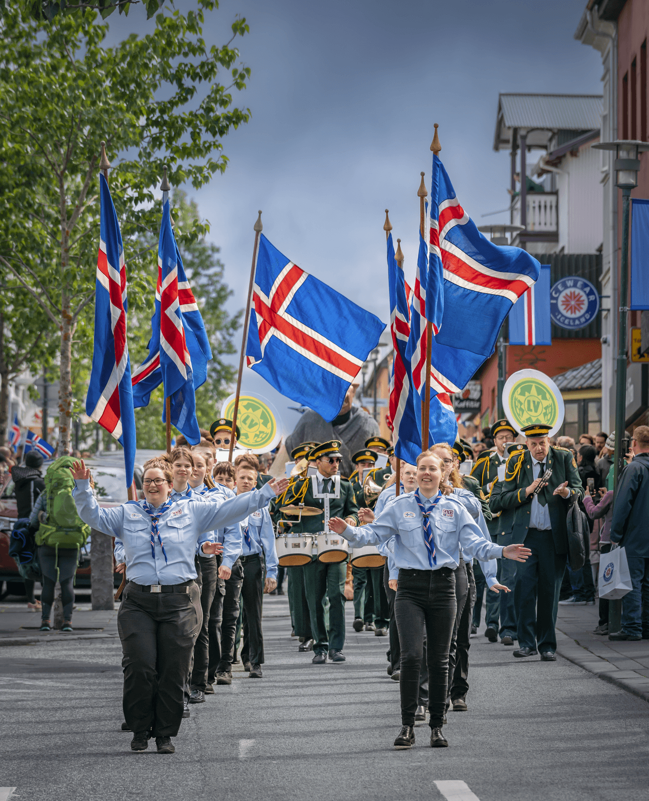 Icelandic scouts lead a festive street parade down a town street during the June 17 National Day celebrations. They wear light blue uniform shirts and dark trousers, waving their arms while marchers behind them carry several large Icelandic flags and a brass marching band follows closely.