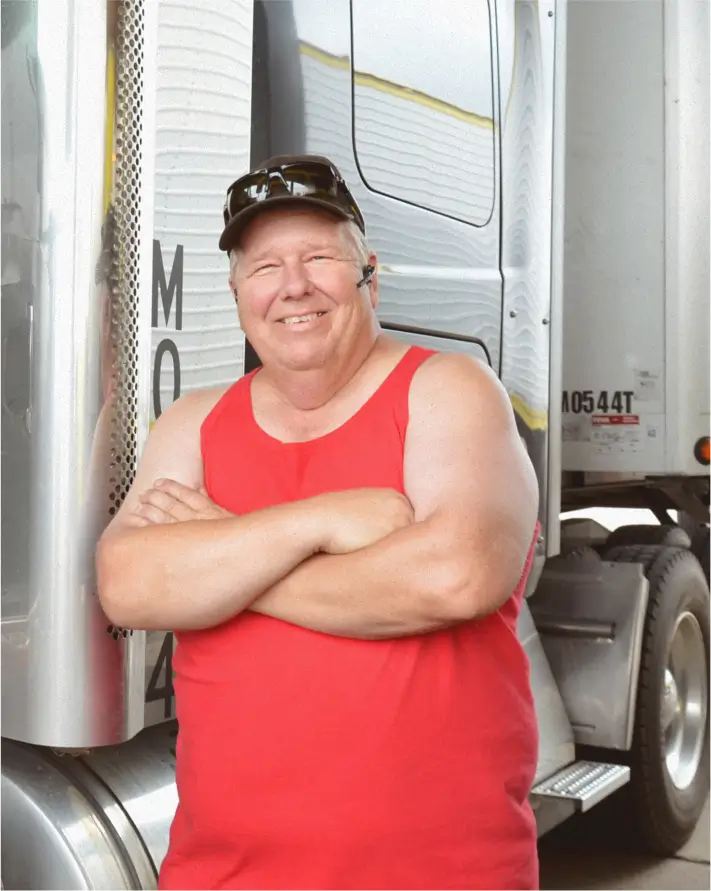A male truck driver in a red shirt leaning against his trailer, arms crossed and smiling