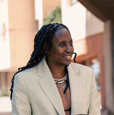 Close-up portrait of a freckled woman with soft pink hair and star earring, natural light, white tank top.