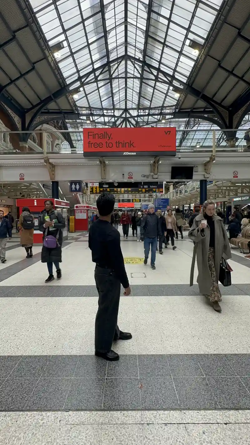 A V7 ‘Finally, free to think’ billboard above the concourse at London Liverpool Street Station, with Christian Harinarain standing among commuters looking up at it