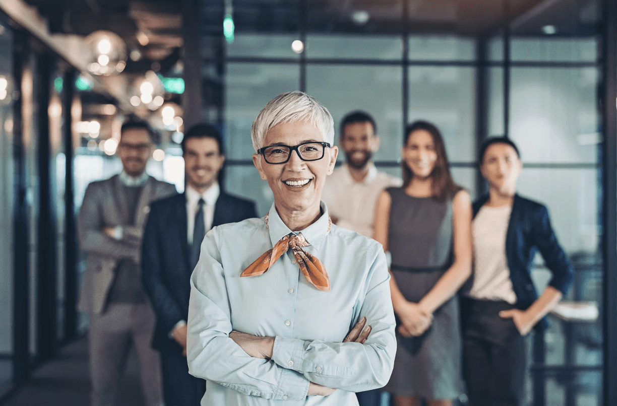 A group of diverse professionals, including a smiling older woman in the foreground, suggesting a successful business or work environment