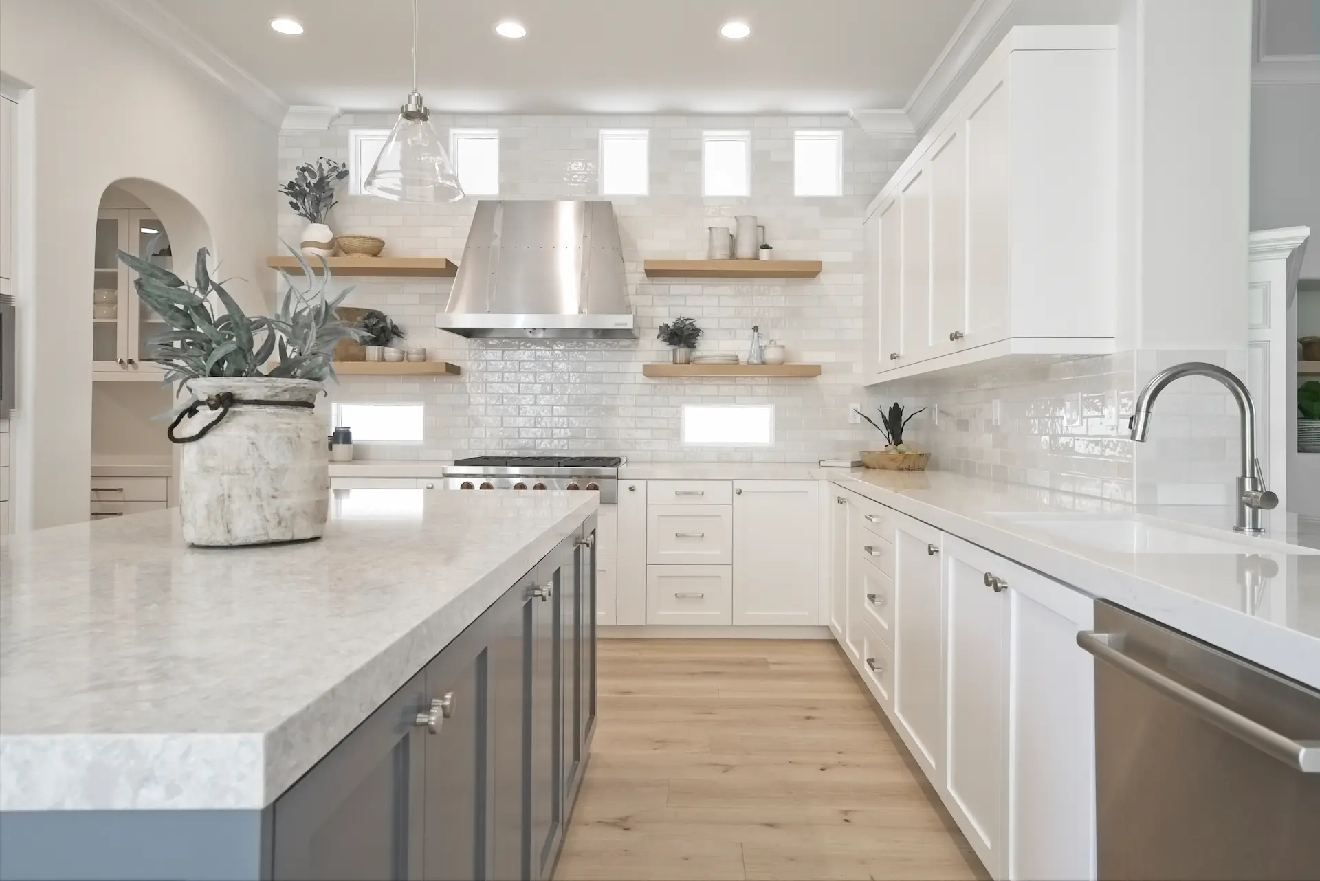 Bright kitchen with white shaker cabinets, floating wooden shelves, and stainless steel hood in Bonita Canyon Remodel.