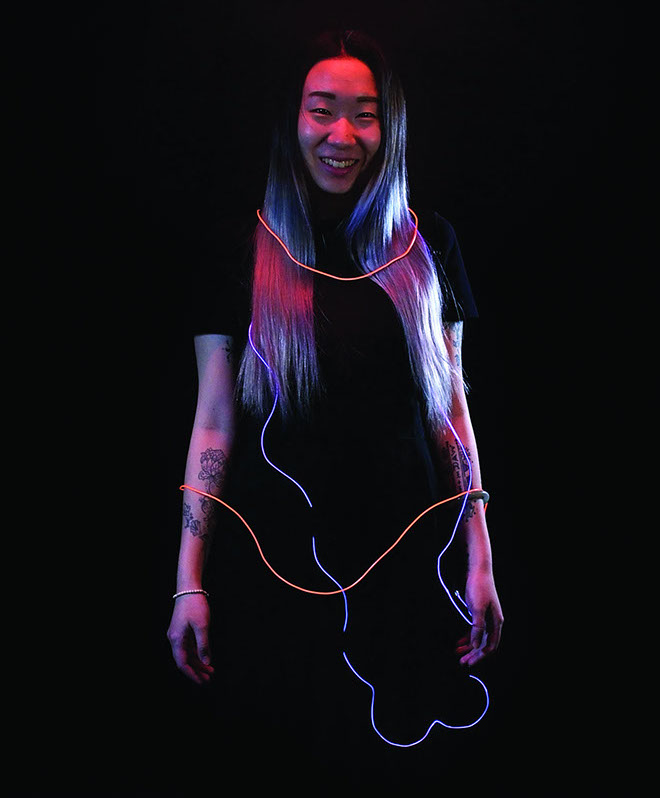 A person with long gray-blue hair stands against a black background, wearing a black short-sleeved top. Their arms are slightly bent and positioned in front of them. A glowing, neon-orange line runs horizontally across their chest and upper arms, resembling a necklace or decorative trim. The person is smiling and looking directly at the camera.
