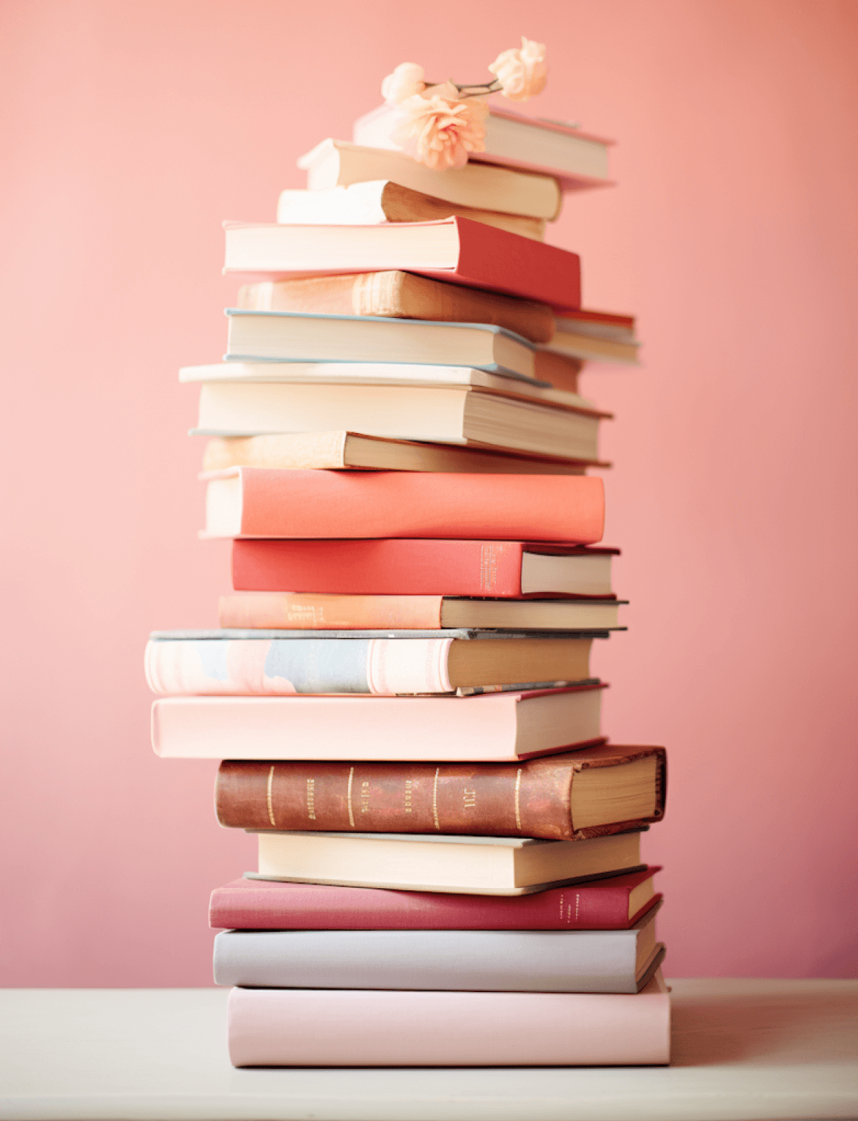 A tall stack of variously colored hardcover books is arranged neatly against a soft pink background, with delicate pink flowers delicately placed on top, creating an inviting and artistic display.