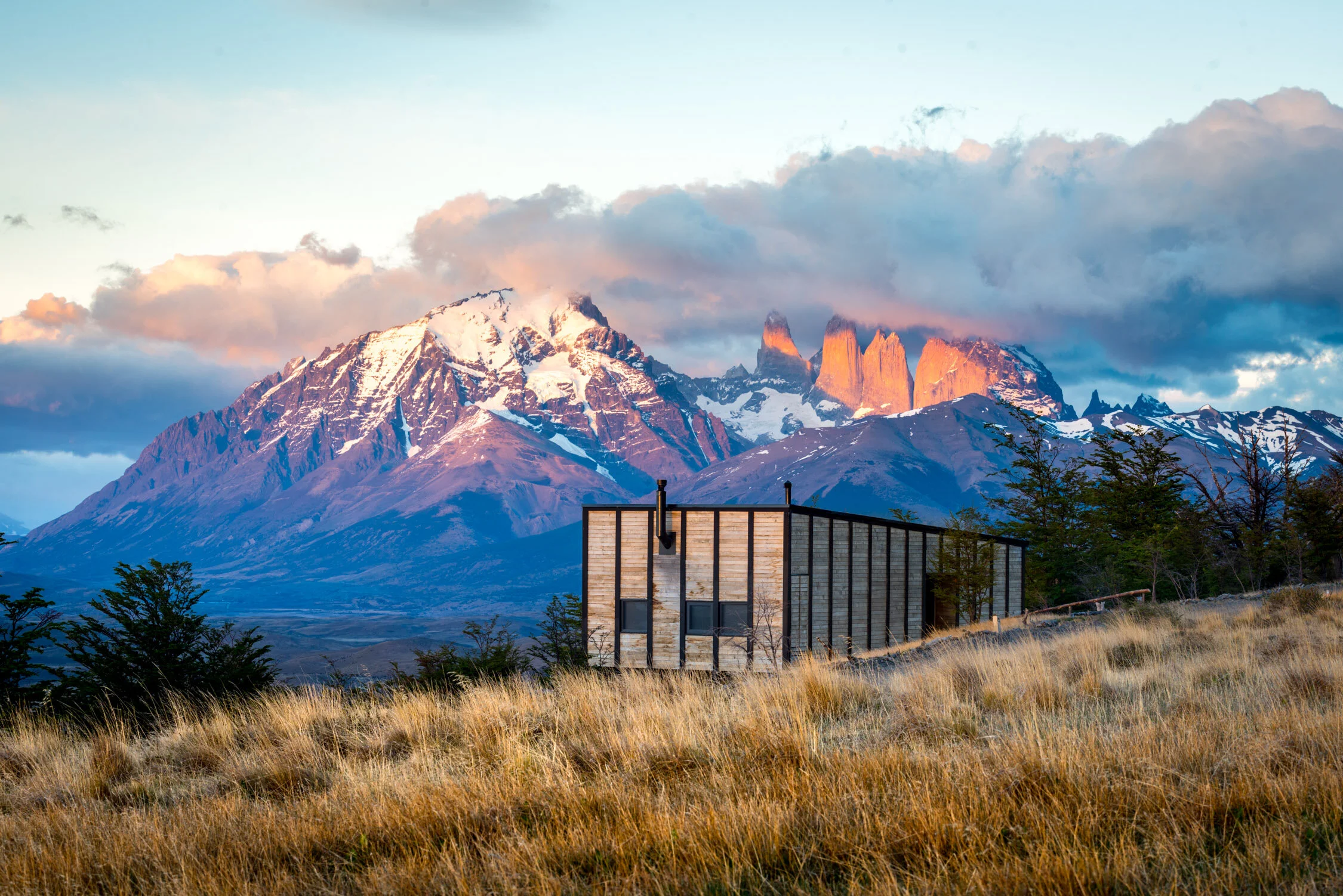 A modern wooden cabin with large glass windows sits in a grassy field before snow-capped mountains and blue skies.