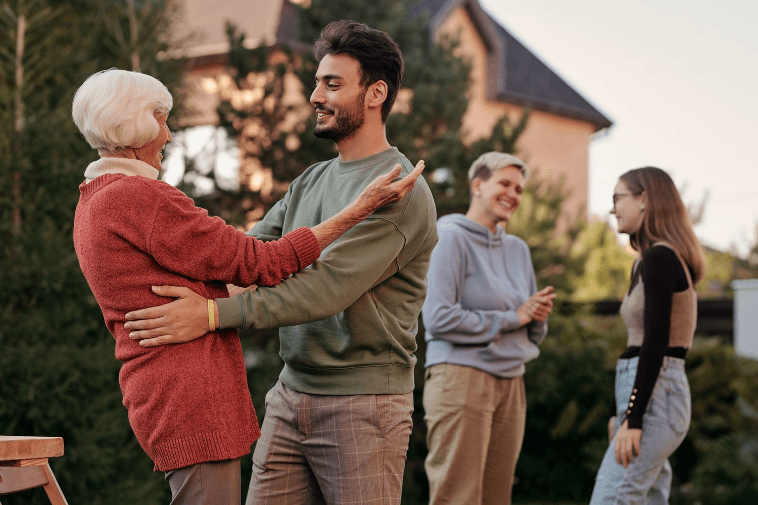 A grandmother greets her grandson in the garden while the rest of the family talks nearby.