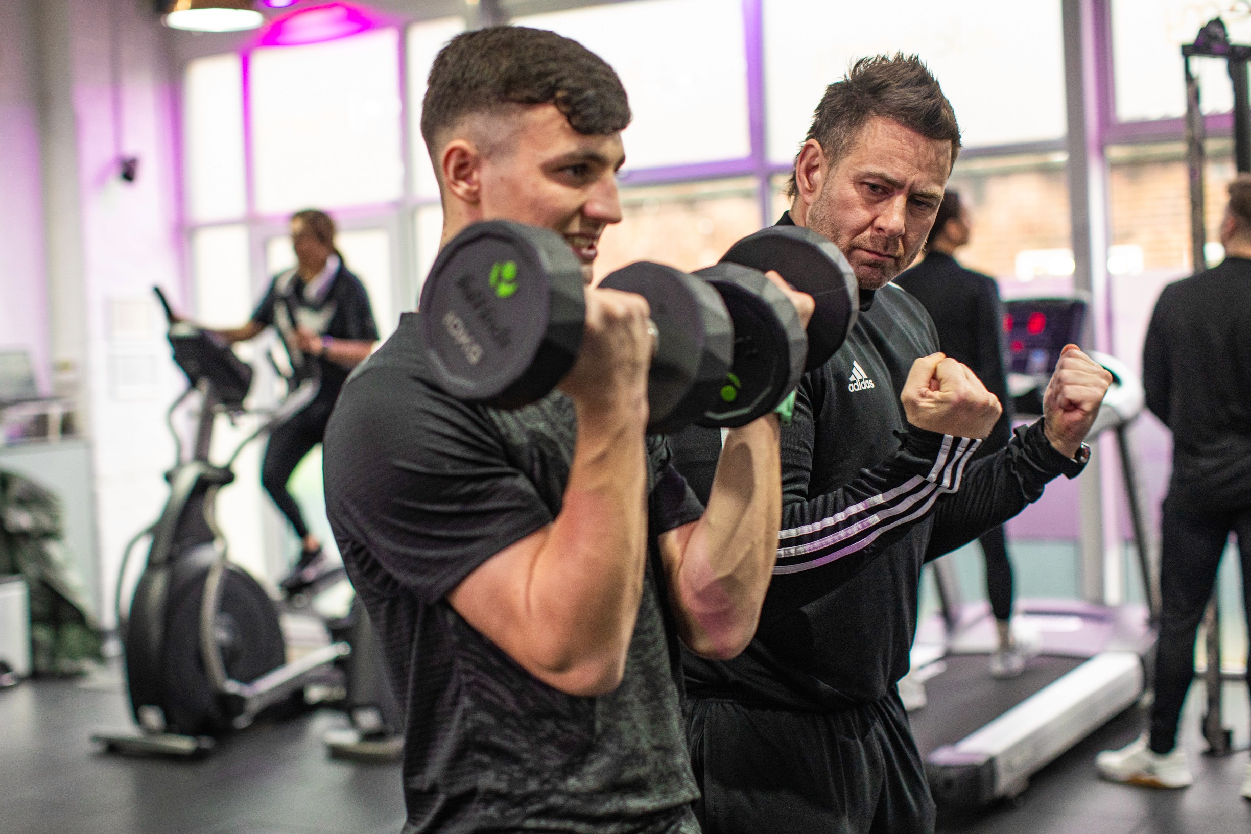 A man and a personal trainer lift dumbbells in a gym, focusing intensely. People in the background use exercise machines, creating an energetic atmosphere.
