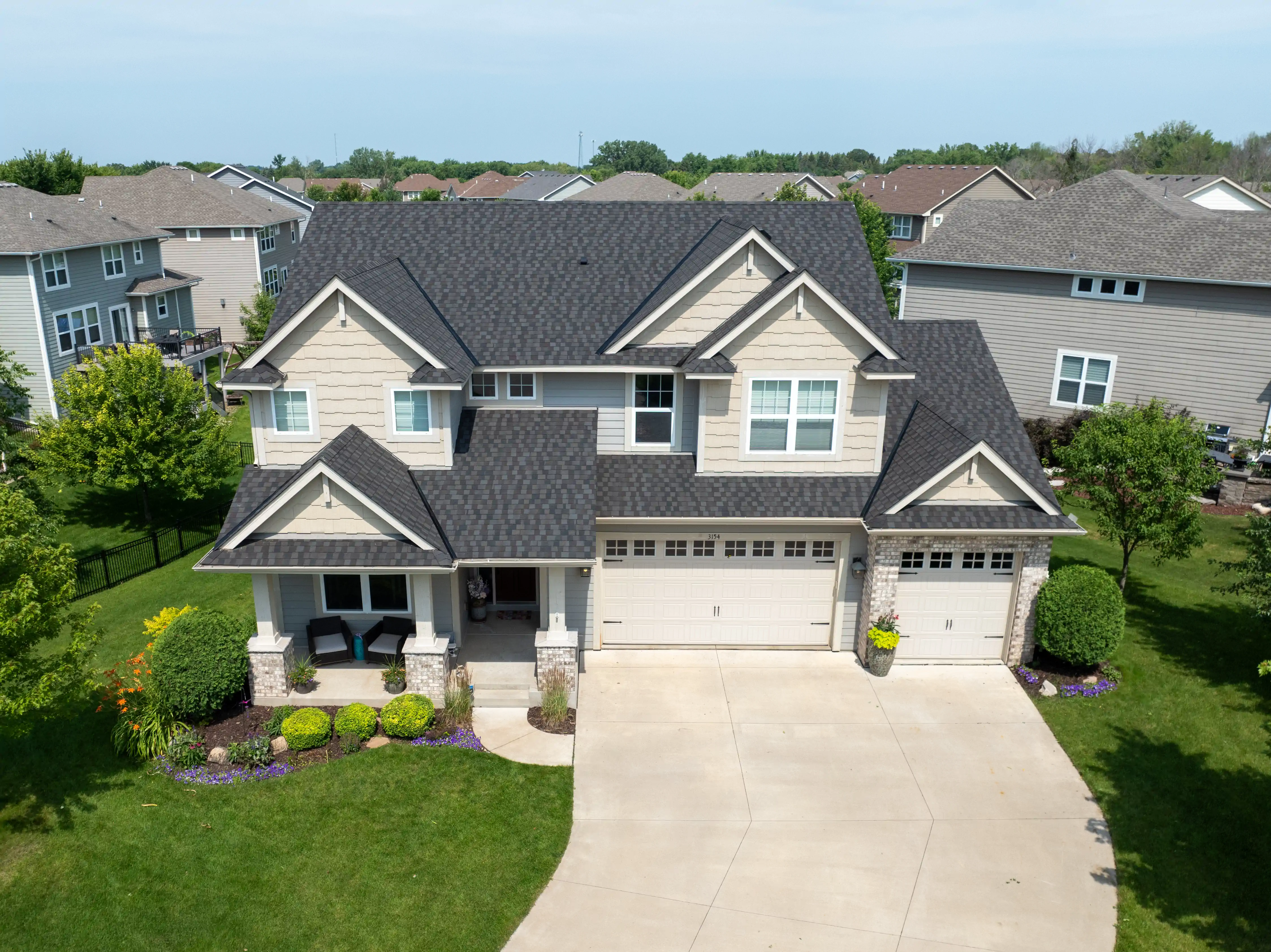 Aerial view of a suburban two-story house with a dark gray roof, beige siding, and a well-maintained front lawn, featuring a driveway leading to a double garage amidst a neighborhood of similar homes, under a clear blue sky.