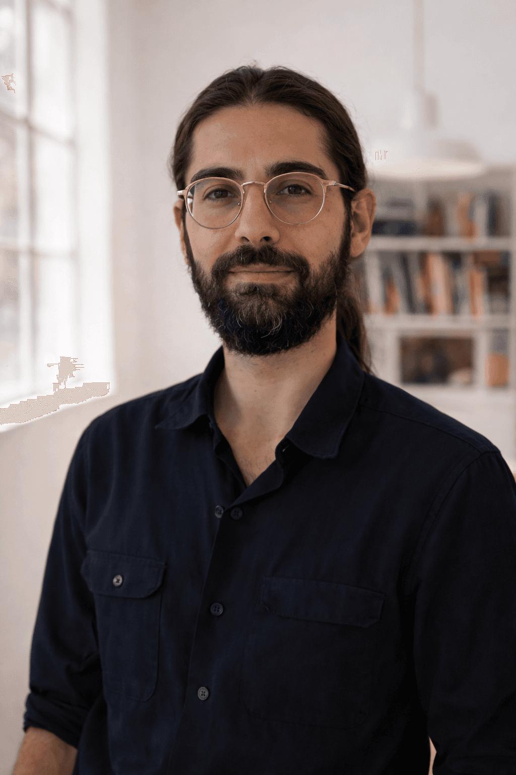A man with glasses and a beard stands by a window, framed by soft sunlight streaming into the room.