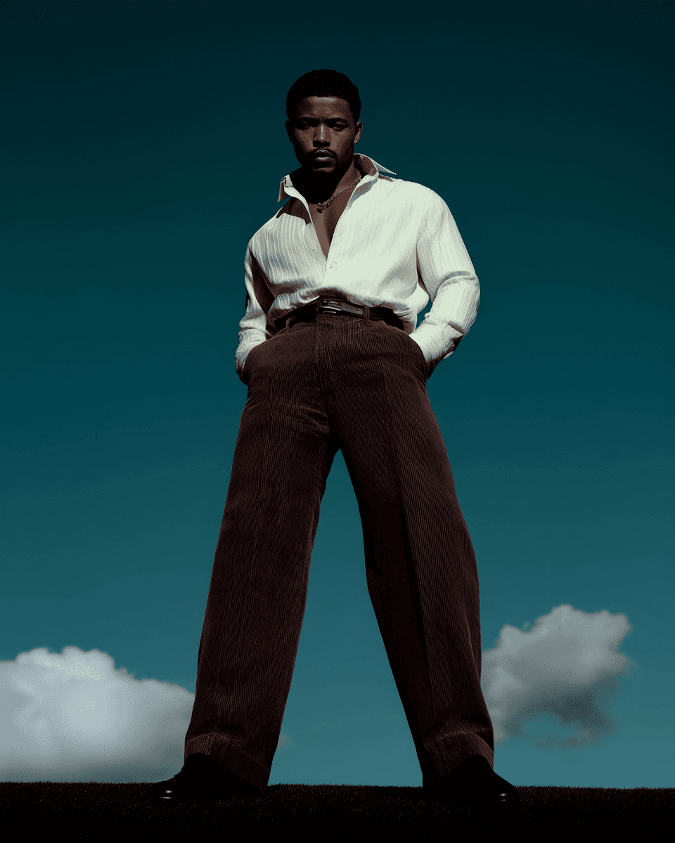 Low-angle shot of a man in a white shirt and high-waisted brown trousers against a deep teal sky with soft clouds.