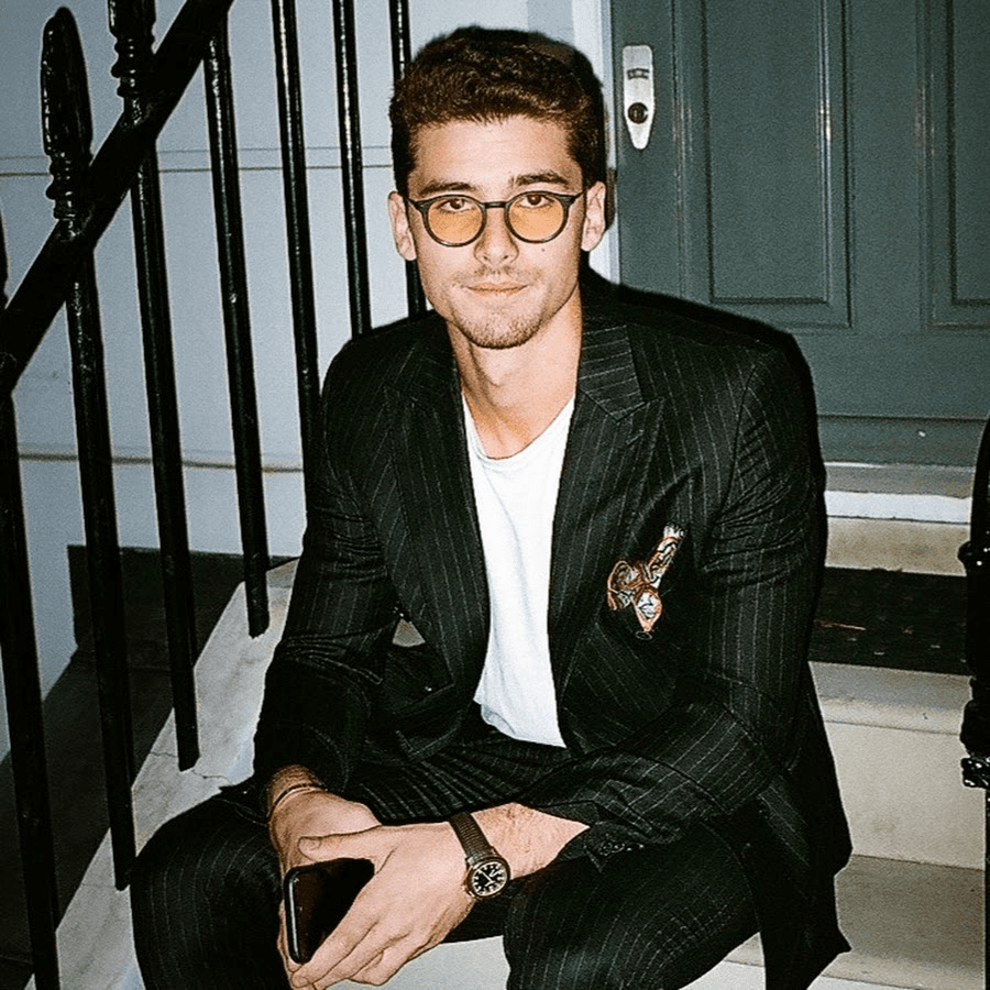 A young man with light brown hair is wearing a black suit jacket and white shirt, standing in a softly lit office setting, conveying professionalism.