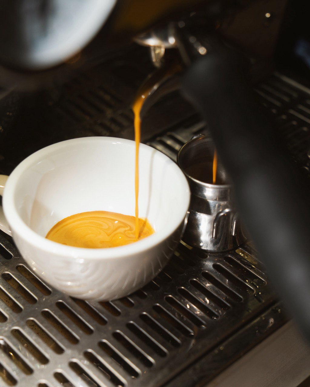 A rich, creamy espresso is being freshly brewed into a white ceramic cup on a stainless steel espresso machine tray, with a small metal milk frothing pitcher placed beside it.