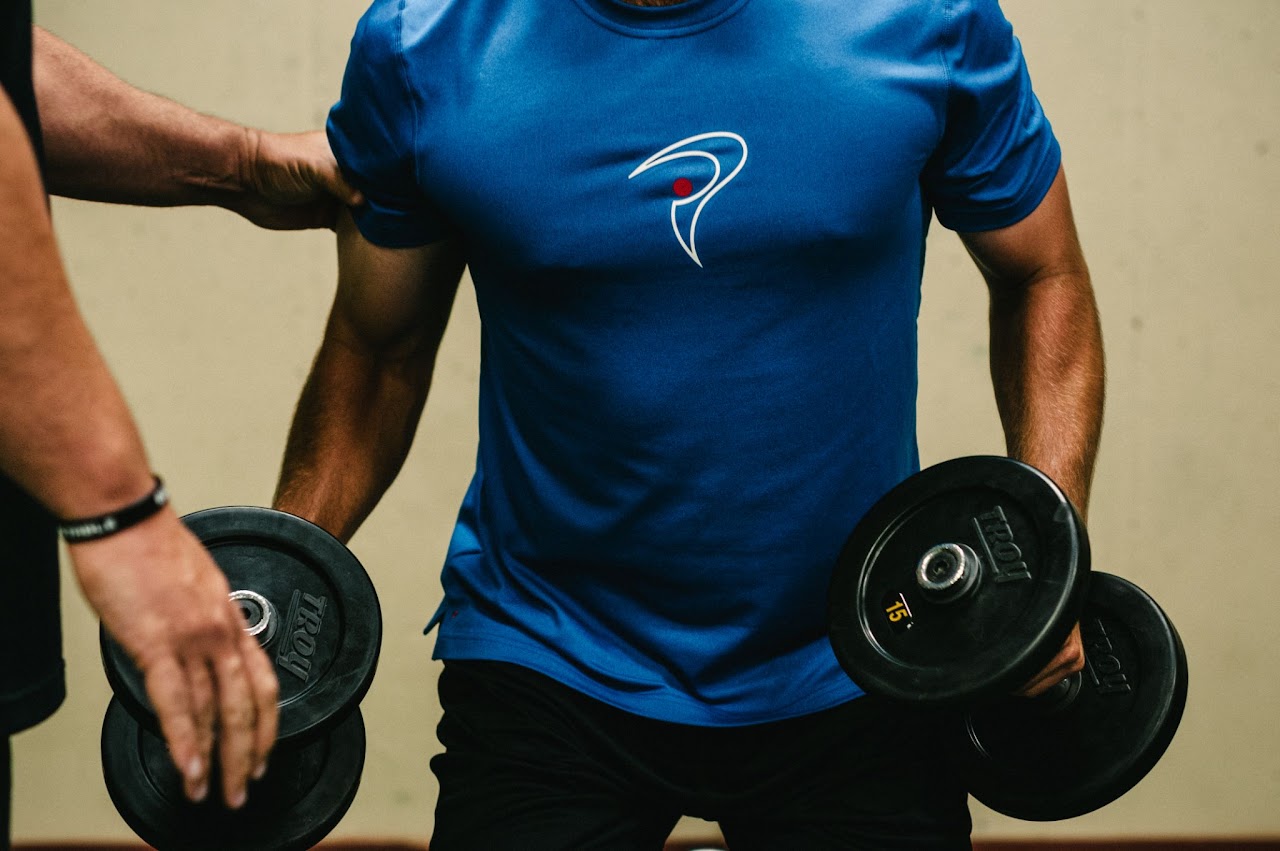A man with a blue shirt lifting dumbbells while being observed by a trainer.