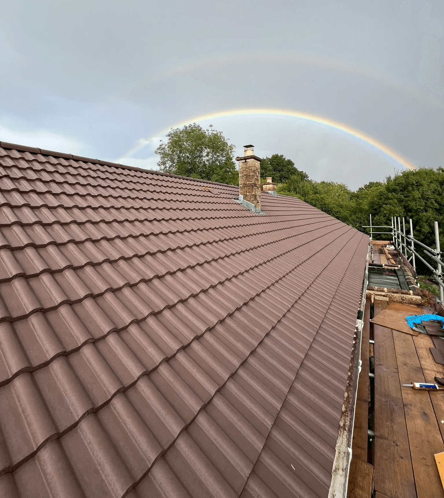 New brown roof with a chimney and a beautiful rainbow arching across the cloudy sky.