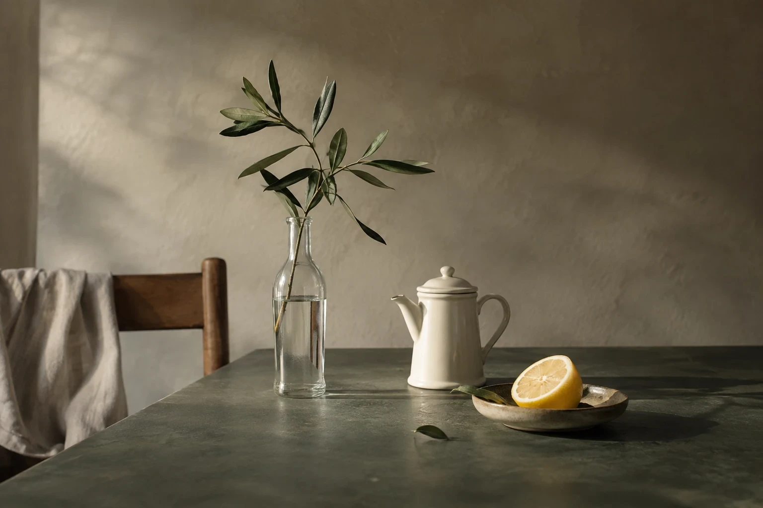 A slim clear bottle with one leafy stem, small ivory coffee pot and cut lemon on dark sage tabletop