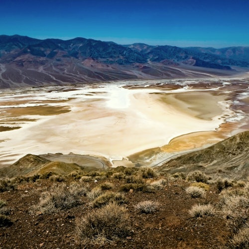 Arid landscape with salt flats stretching across the foreground, surrounded by dry hills and distant mountains under a clear blue sky.