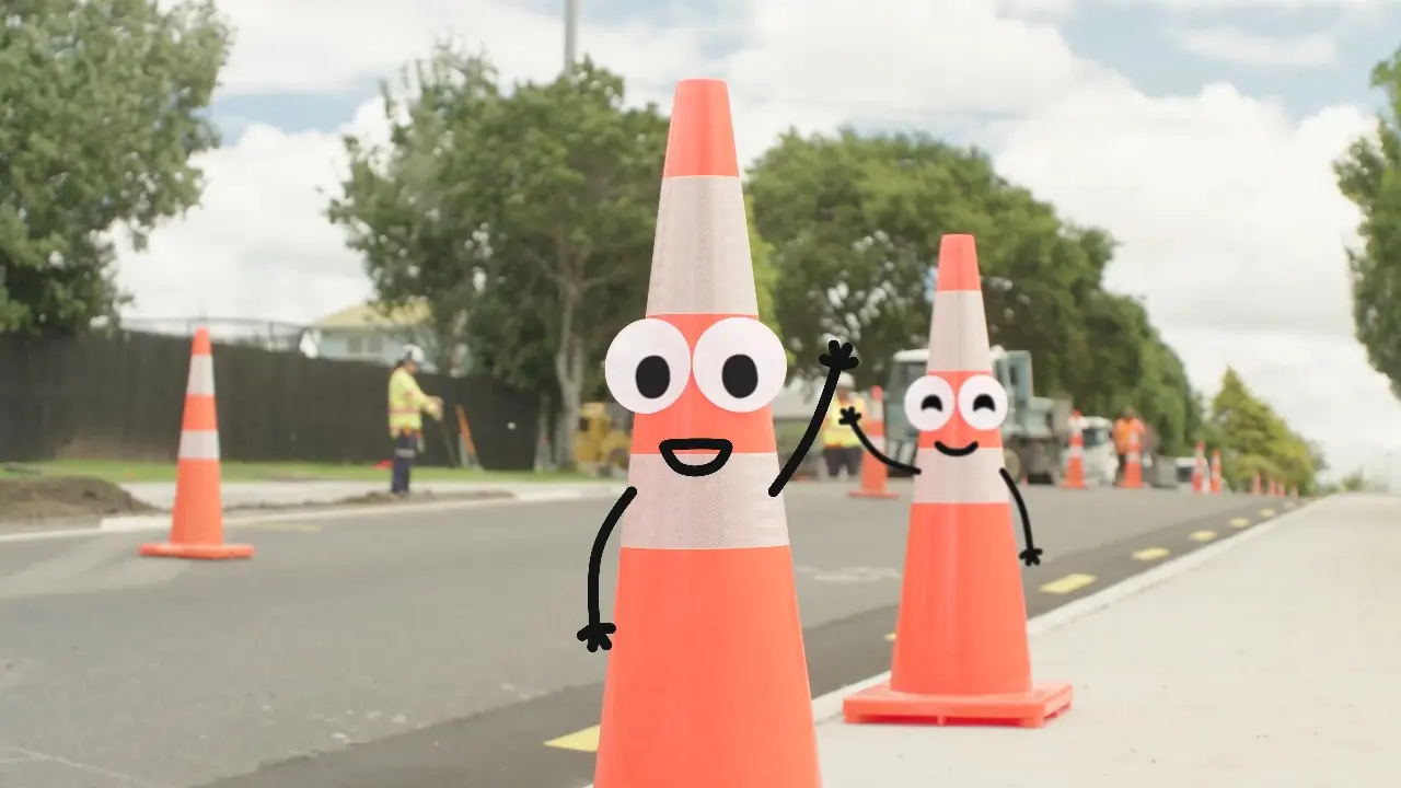 Two road cones with googly eyes, drawn smiles and stick arms wave beside a road