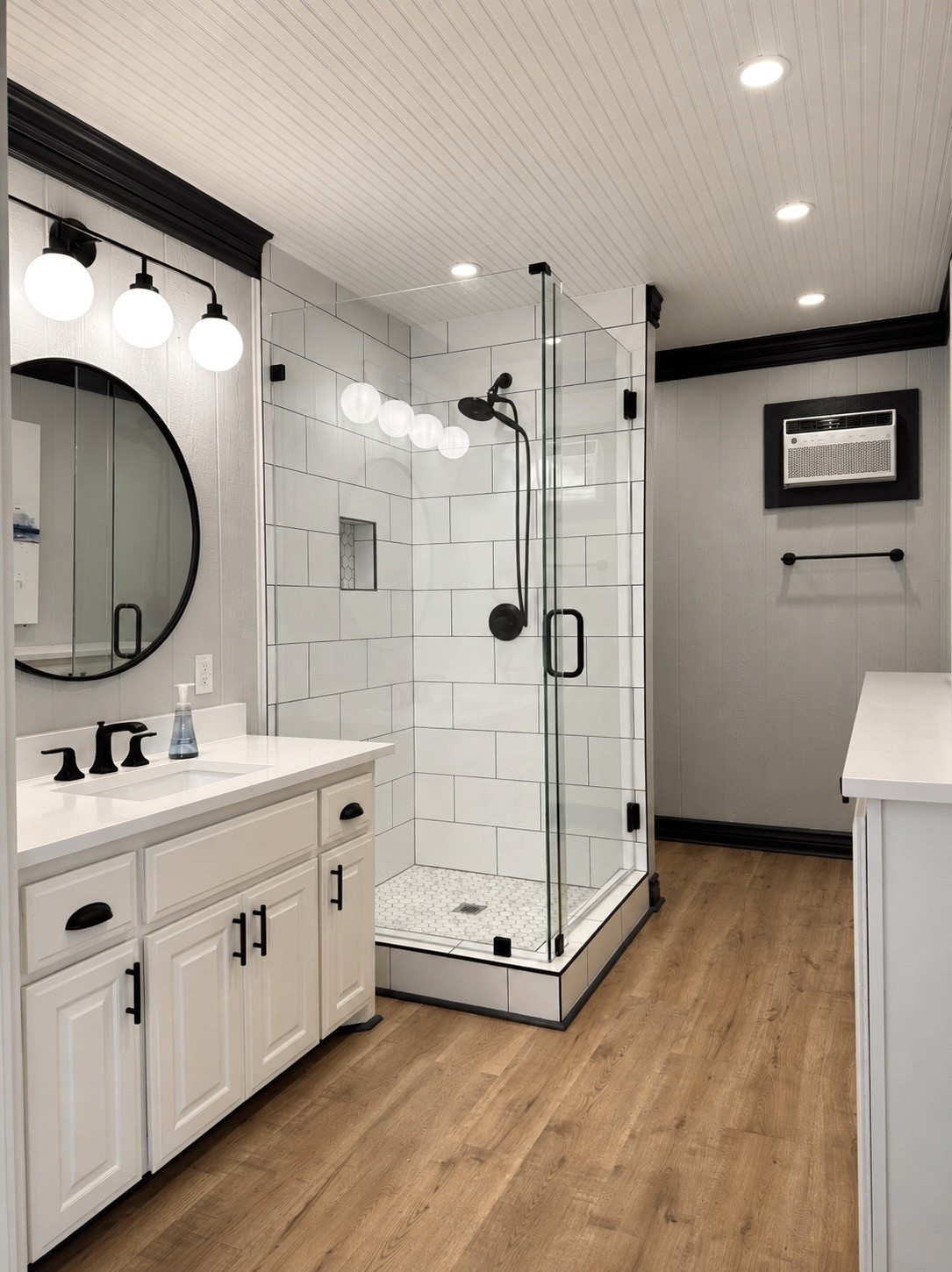 A modern bathroom features a spacious glass-enclosed shower with white subway tiles, a wooden floor, a round mirror above a white vanity sink with dark fixtures, and overhead recessed lighting.