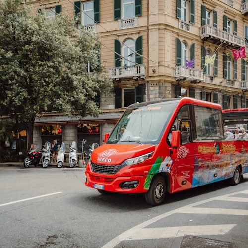 Red tourist bus on a city street with scooters parked near a large building adorned with balconies and colorful butterfly decorations.