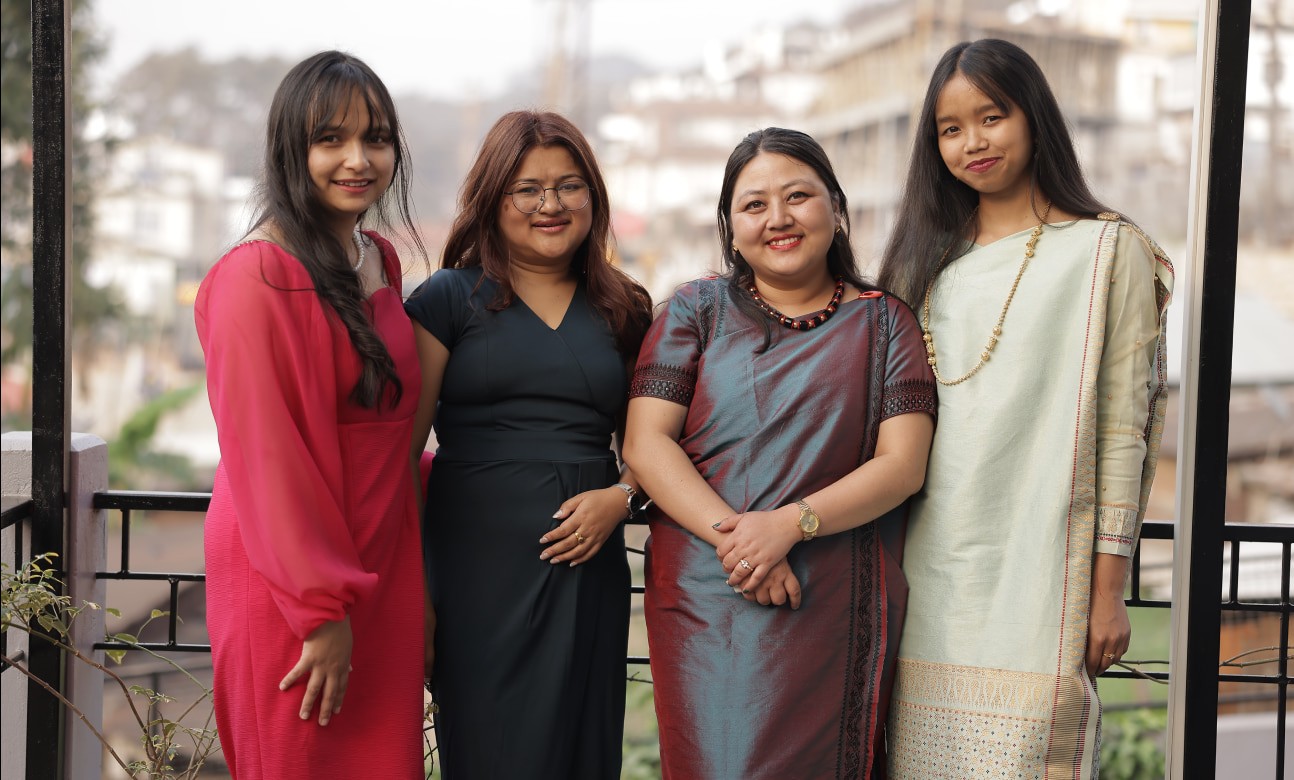 Four women smiling at the camera, wearing different colored dresses and shawls.