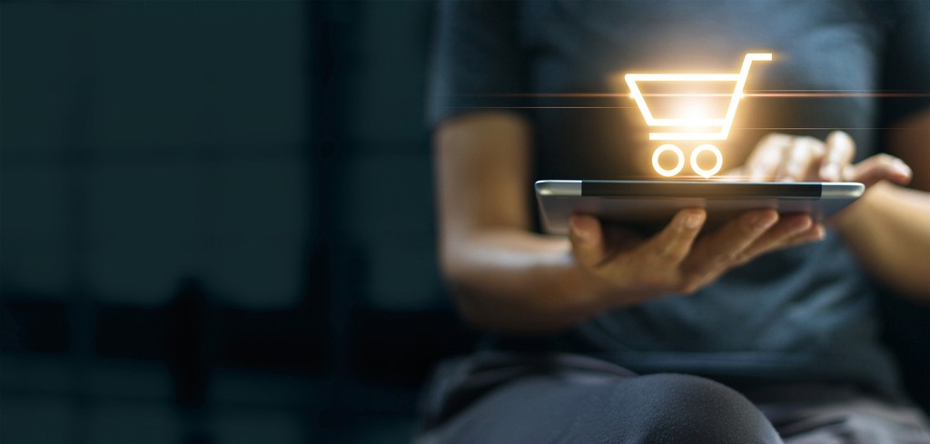 Pessoa segurando tablet com ícone holográfico de carrinho de compras iluminado, representando e-commerce mobile e experiência de compra digital pelo dispositivo móvel - FindUP - Tendências de TI para o varejo