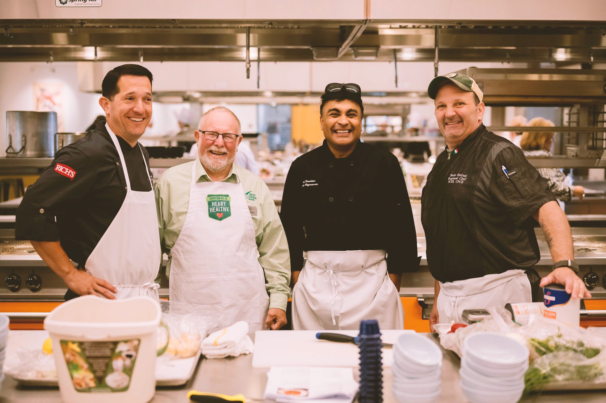Person with a mustache wearing a black t-shirt carrying a tall stack of pizza boxes inside a dimly lit bar or restaurant.