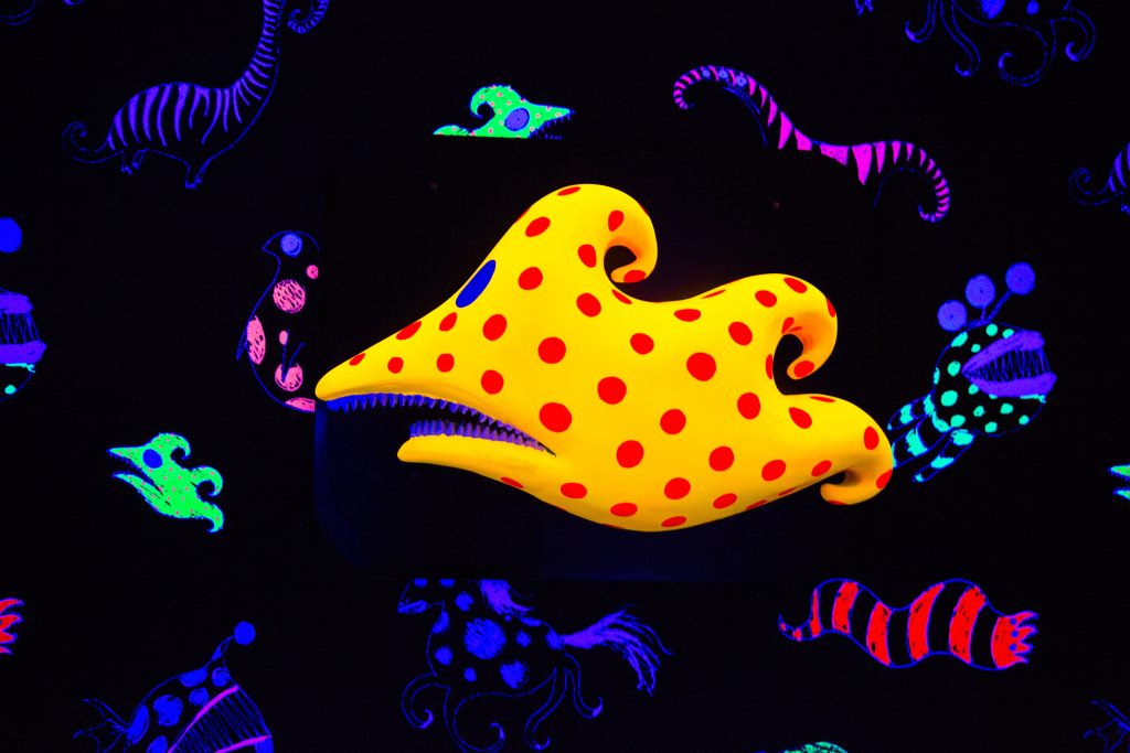 Gelbe Fischskulptur mit roten Punkten zwischen leuchtenden Schwarzlichtfiguren, Beispiel für 3D-Gestaltung